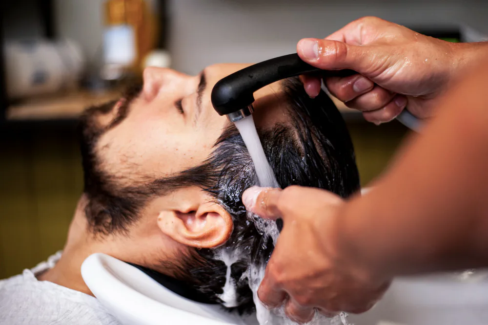 Manos de un profesional lavando con agua y espuma el cabello de un hombre tumbado en un lavacabezas