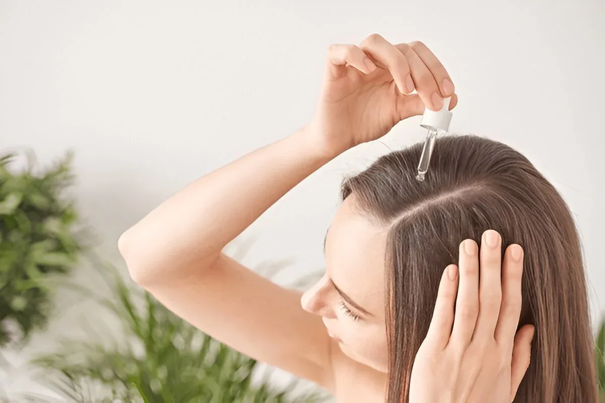 Mujer aplicándose un producto con cuentagotas en la raíz del cabello, con la otra mano sujetando el pelo